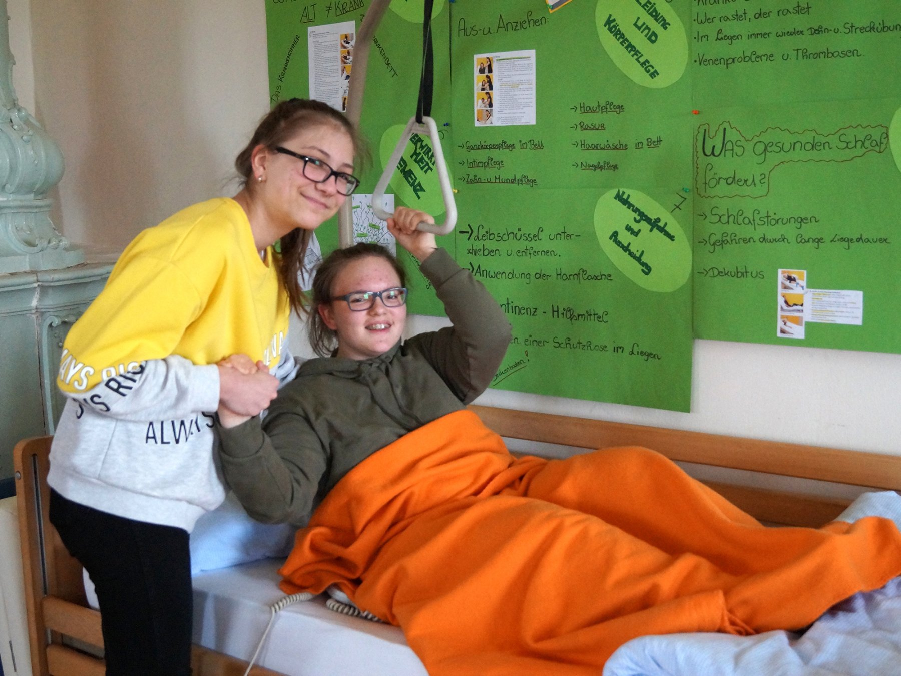 Schülerin stützt eine andere Schülerin, die in einem Pflegebett liegt
